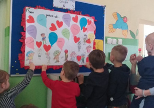 Plastykoterapia w grupie 5. Grupa dzieci zawiesza na tablicy wspólnie zrobiony plakat dot. praw dziecka.